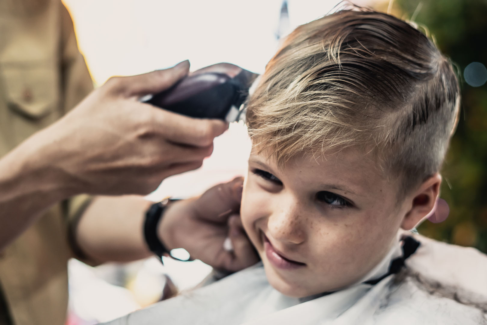 Barber shop blond boy customer. Hairdresser hand electric tool clipper. Child portrait real emotions